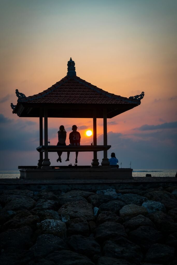Silhouettes of people sitting in a pagoda overlooking a stunning sunset at Kuta Beach, Bali.